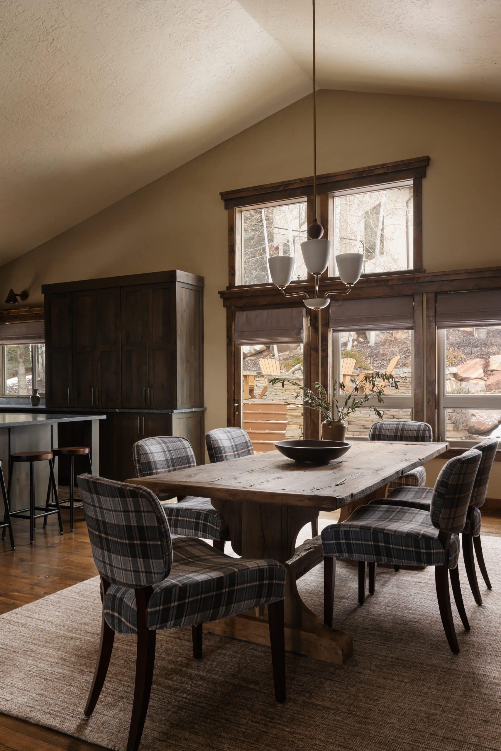 Dining area with a plank table, plaid armchairs, and windows framing the mountain landscape