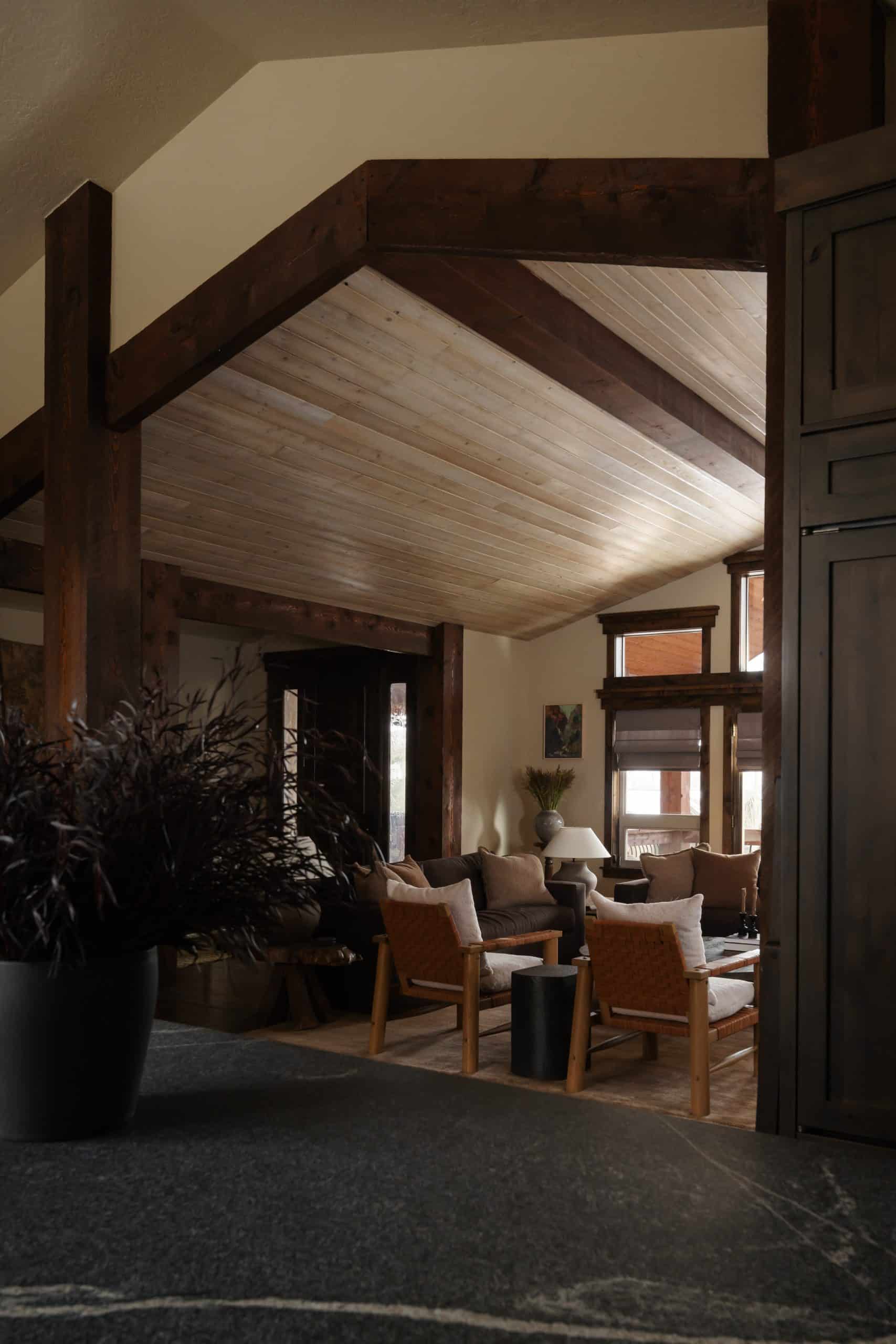 View through timber-framed openings into the living room with vaulted wood ceiling and woven-leather lounge chairs