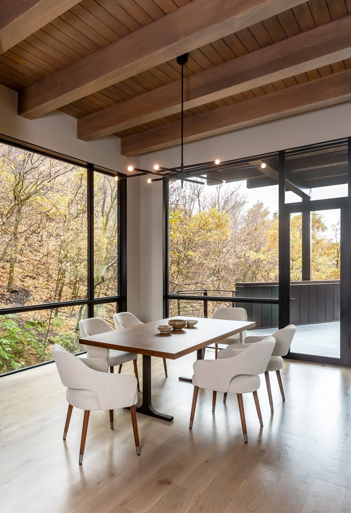 Dining area with a walnut table and cream chairs framed by corner windows onto the autumn woods