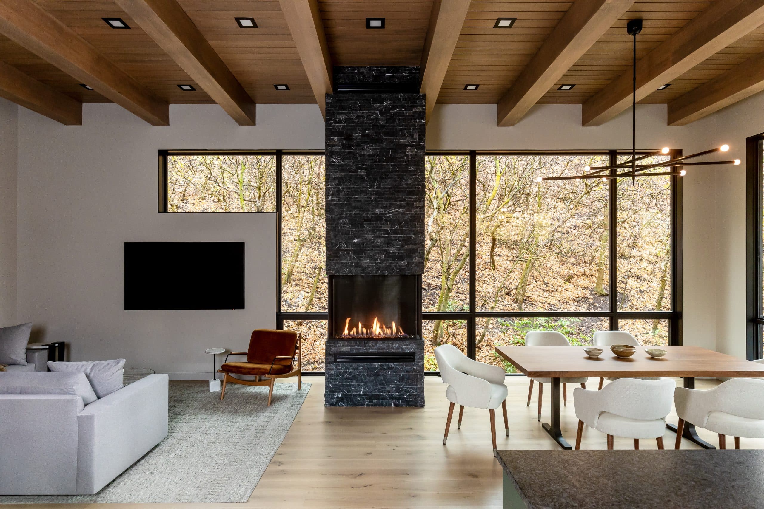 Great room with exposed wood beams, black stacked-stone fireplace, and floor-to-ceiling forest views