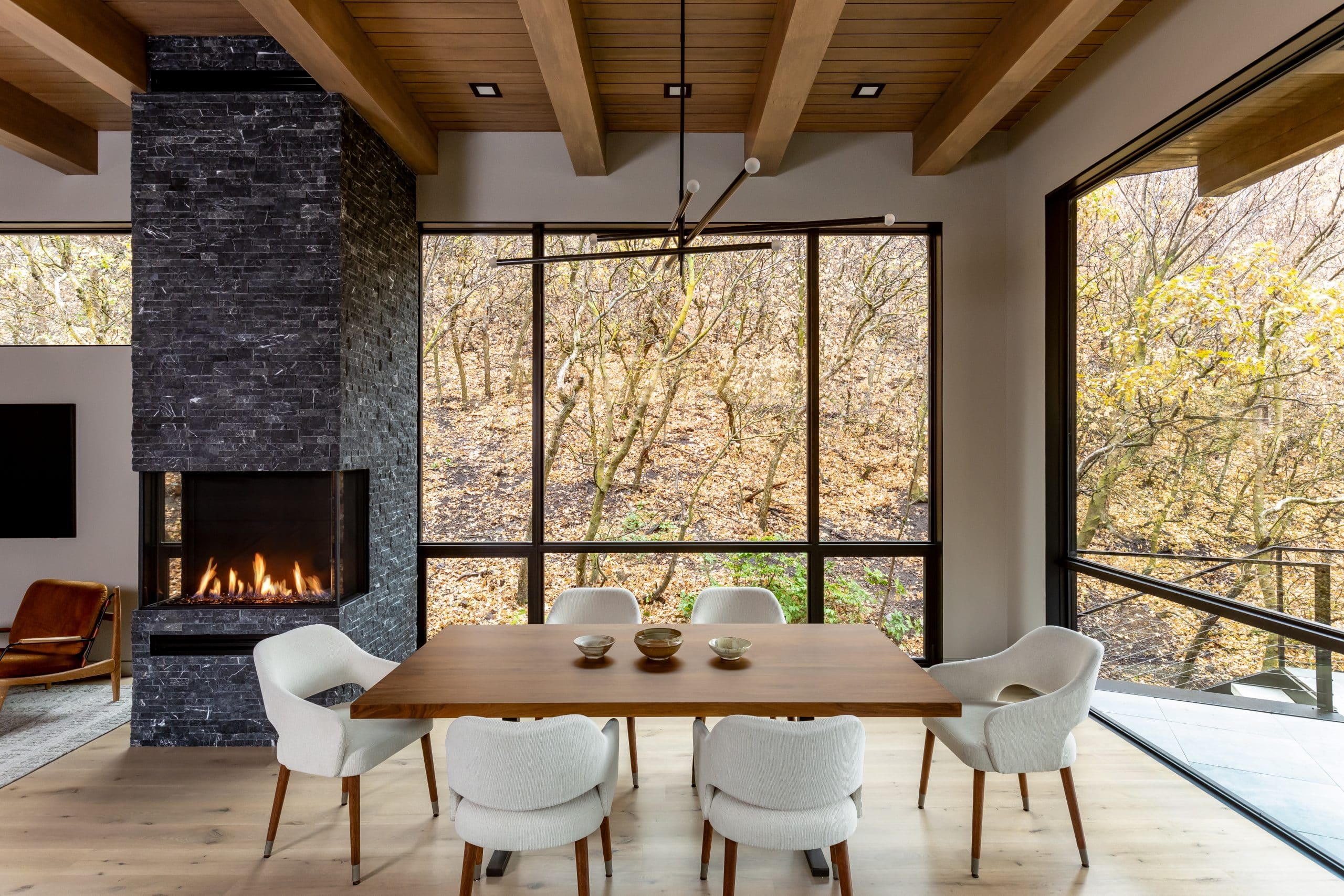 Dining table and upholstered chairs beside the black stone fireplace and window wall