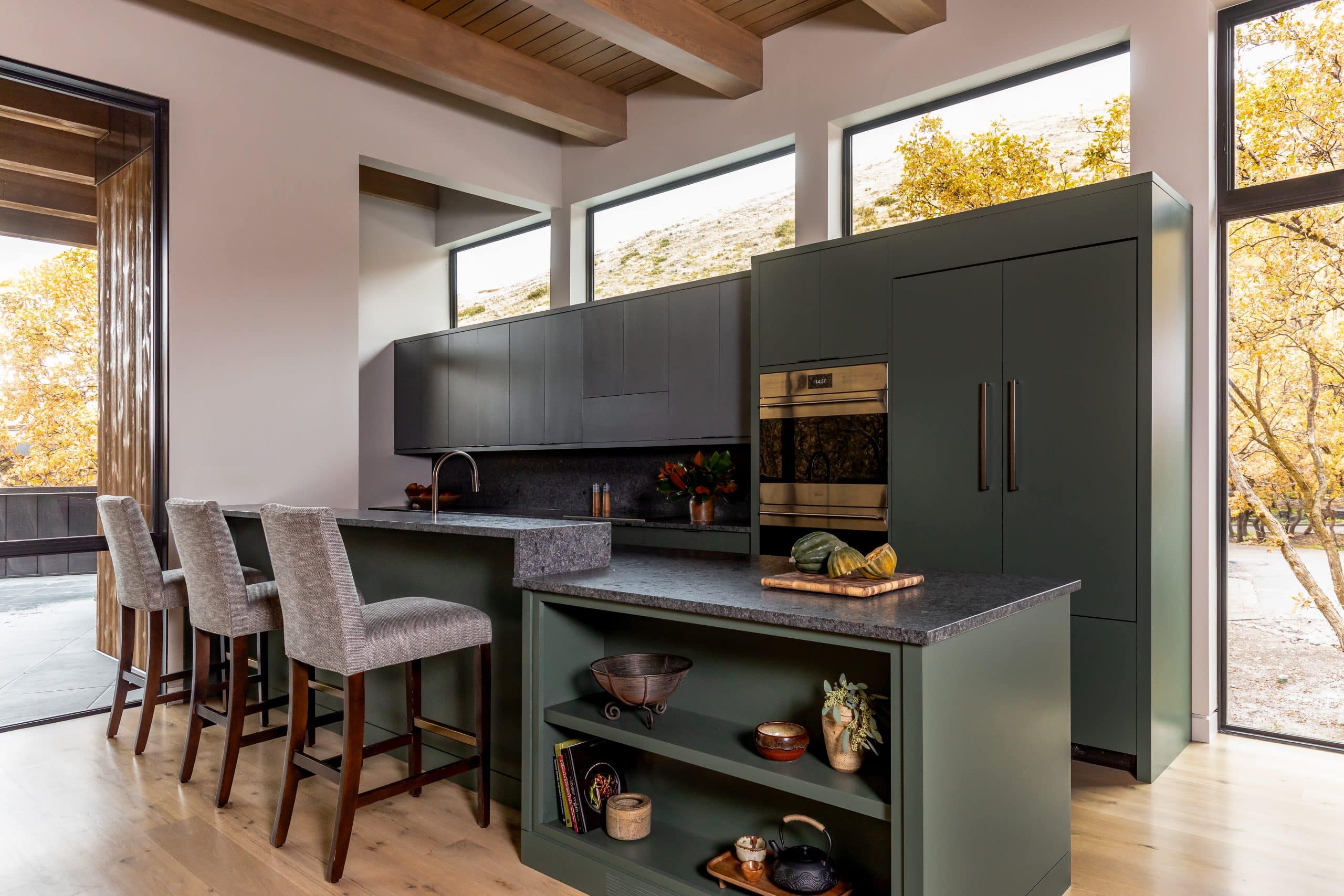 Green kitchen with a stone-topped island, open shelving, brass wall oven, and counter stools