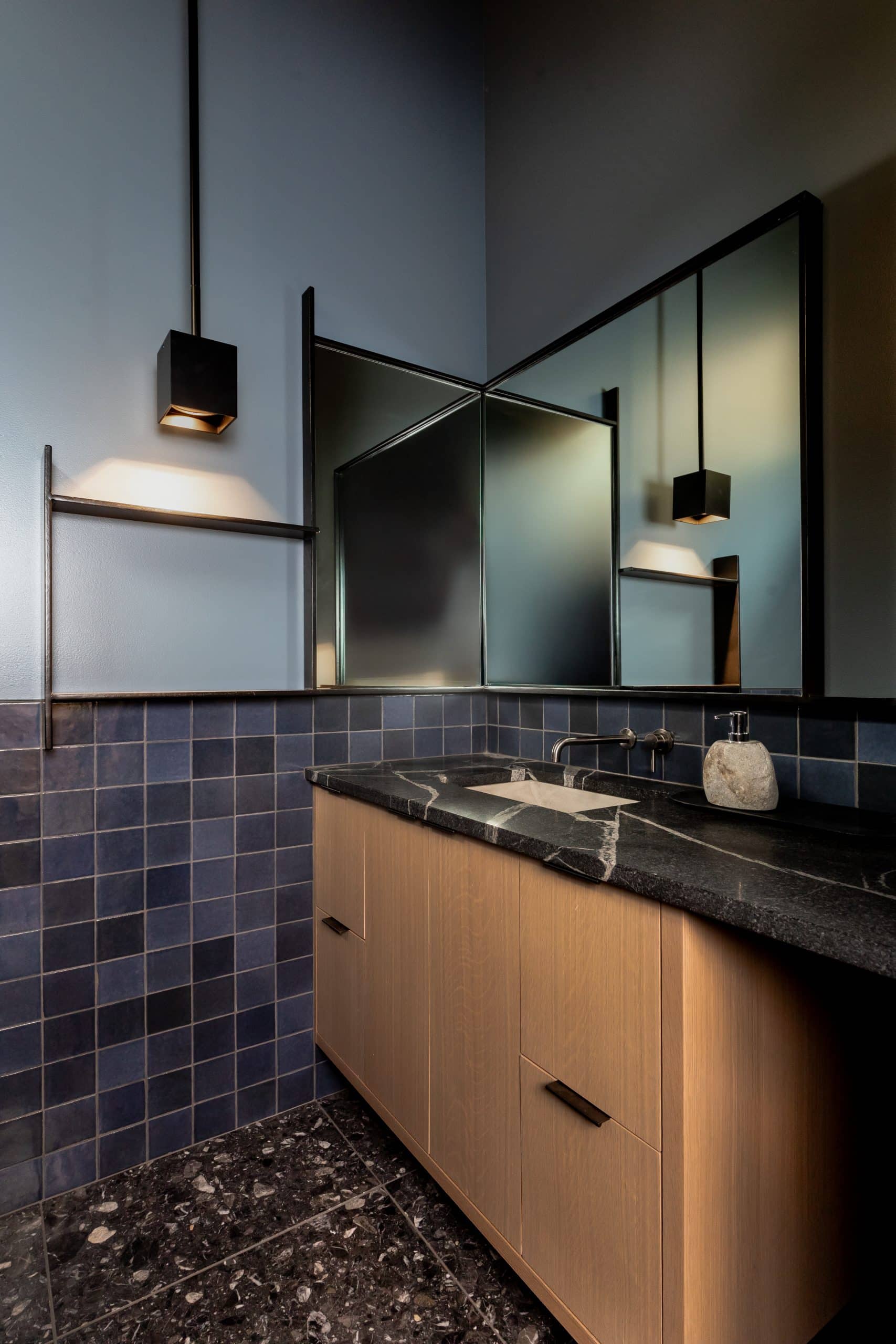 Powder room with blue tile, corner mirrors, an oak floating vanity, and dark terrazzo floor