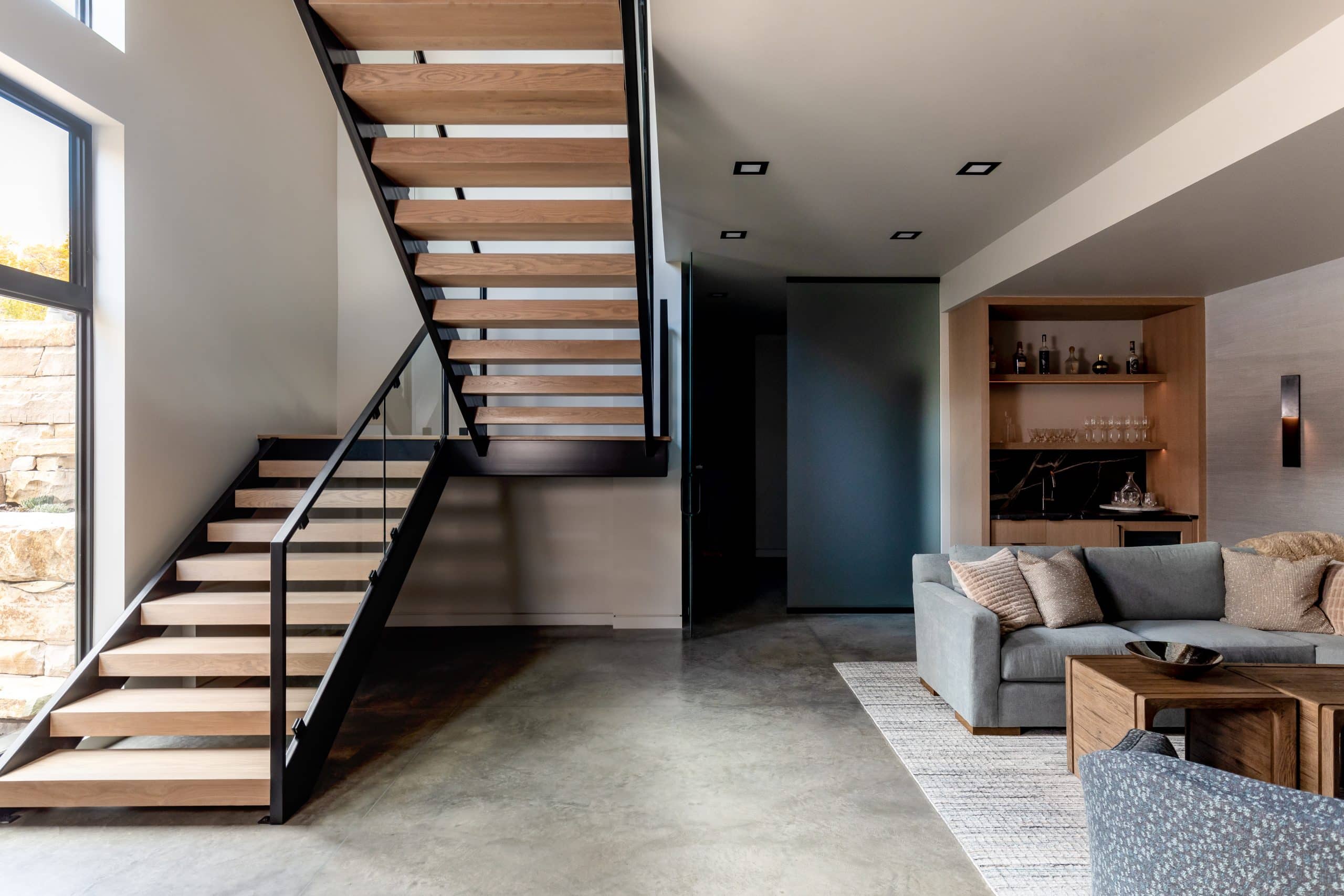 Open-tread staircase over a polished concrete floor with a wet bar and gray sofa beyond