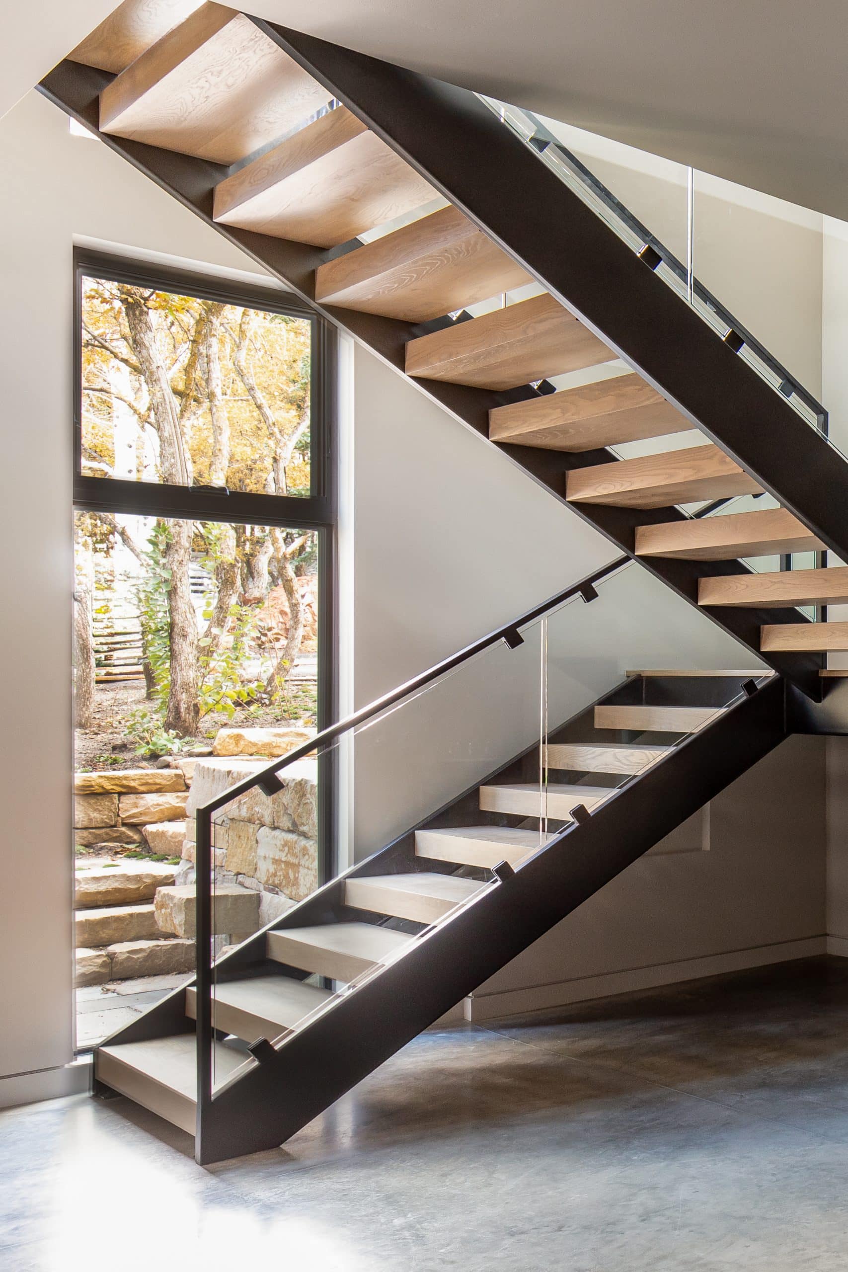 Floating oak-and-steel staircase beside a tall window framing a stone retaining wall and trees