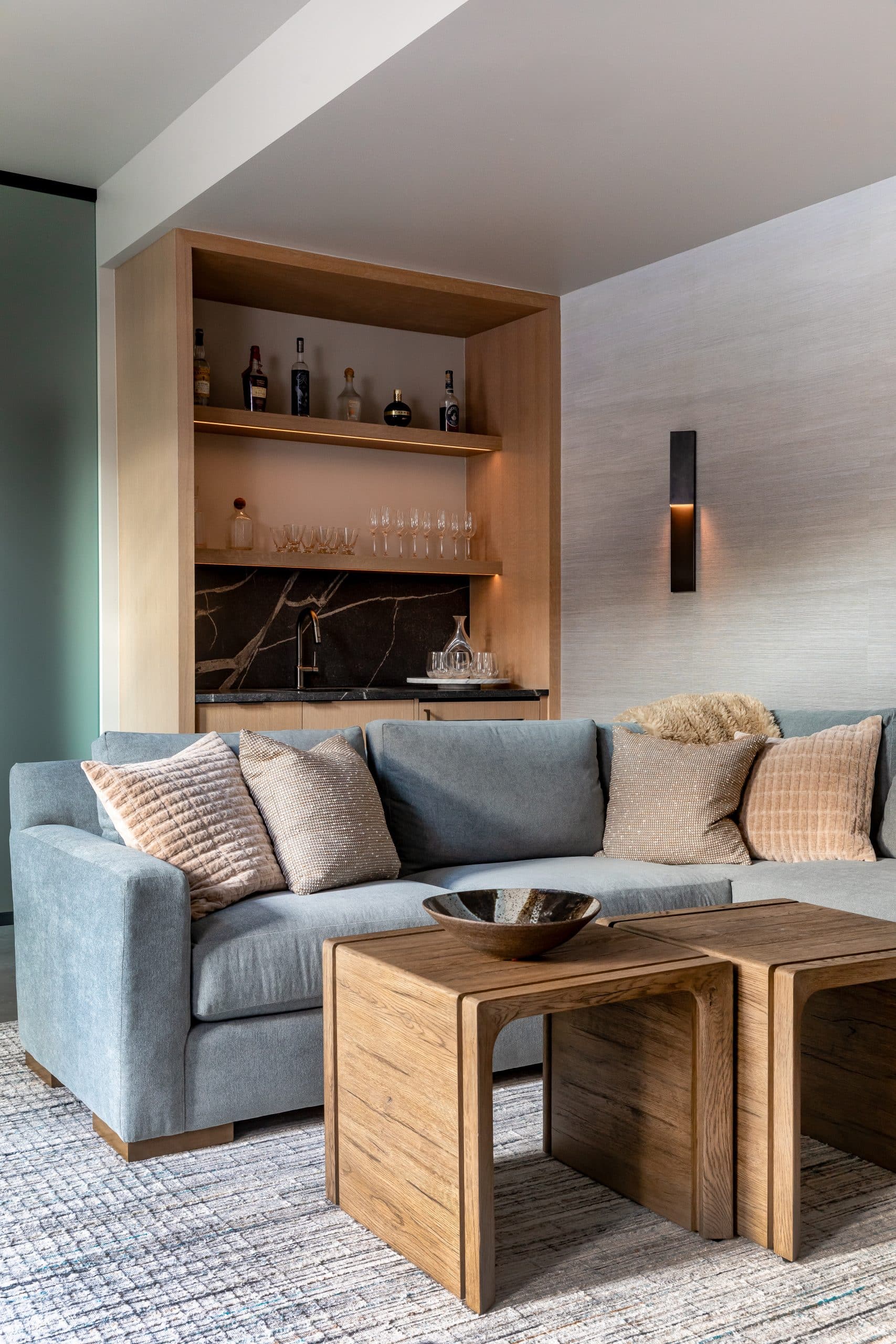 Lower-level seating area with a blue-gray sofa, oak wet bar, and nesting wood tables