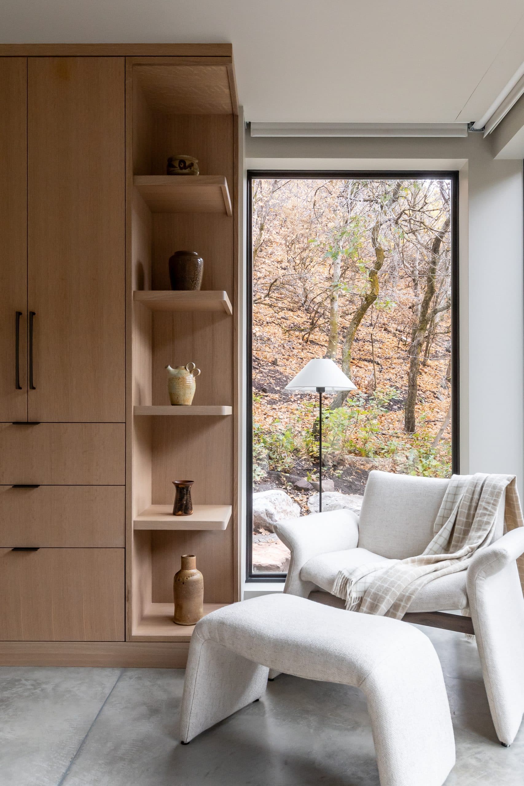 Sitting corner with a boucle chair and ottoman beside oak shelving and a window onto autumn trees