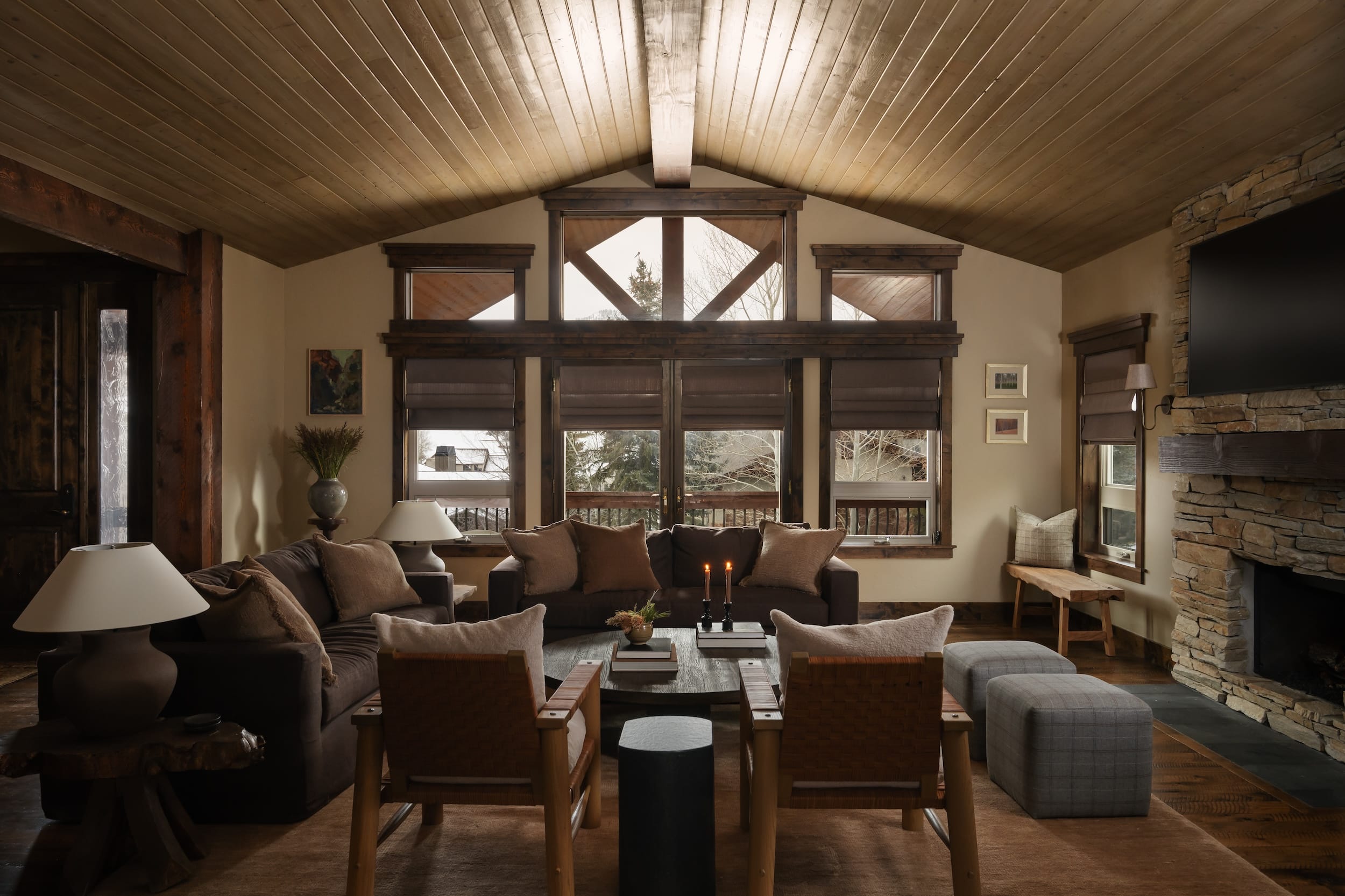 Luxury mountain rustic living room in Park City with sofas, dining table, chairs, and candles, featuring large windows overlooking pine trees.